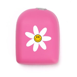 Può includere: Una custodia rosa brillante di forma rettangolare arrotondata con una grafica a margherita bianca e una faccina sorridente gialla al centro. La custodia ha una finitura opaca liscia e un piccolo anello in metallo argentato sul bordo.