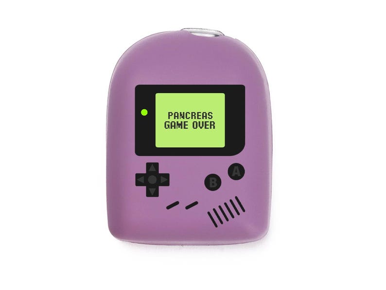 May include: A lavender-coloured novelty item shaped like a retro handheld game console. The screen displays "PANCREAS GAME OVER" in white text on a green background. Black buttons and directional pad are also visible.