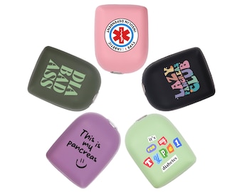 May include: Five protective covers for medical devices in pink, olive green, black, purple, and light green. The pink cover features a red medical symbol and text. The olive green cover says DIA BAD ASS. The black cover says LAZY PANCREAS CLUB. The purple cover says This is my pancreas with a smiley face. The light green cover says it's FOR MY TYPE 1 diabetes.