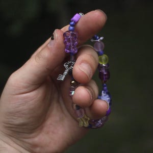 Puede incluir: Una pulsera de cuentas con una variedad de cuentas moradas, amarillas y transparentes, que incluye un dije translúcido con forma de oso morado y un dije de metal plateado con forma de llave, ensartados en un cordón flexible.