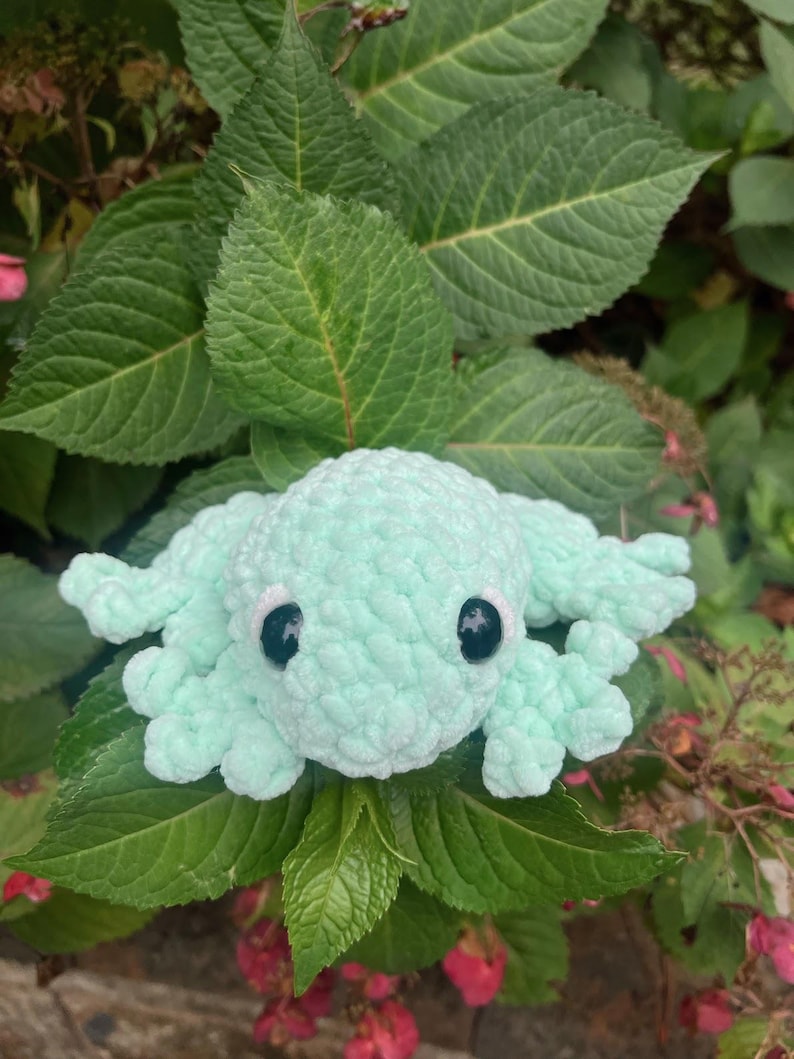 May include: A light blue crocheted octopus with black eyes. The octopus is sitting on a bed of green leaves.