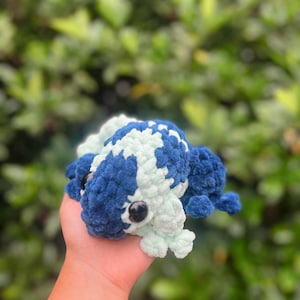 May include: A crocheted frog toy with blue and light green yarn. The frog has black eyes and a textured surface.