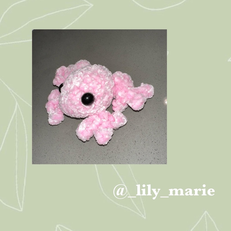 May include: A pink crocheted frog with black eyes. The frog is lying on its belly with its legs outstretched.