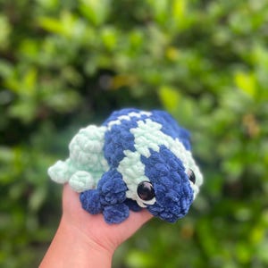 May include: A crocheted frog plush toy with blue and green fur. The frog has large black eyes and a small, upturned mouth.