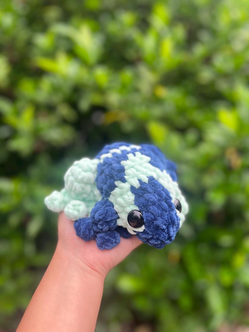 May include: A crocheted frog plush toy with blue and green fur. The frog has large black eyes and a small, upturned mouth.