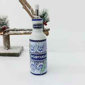 Può includere: Una bottiglia di olio d'oliva in ceramica bianca con un design a motivi blu e bianchi che raffigura pesci e la parola "PORTUGAL". La bottiglia ha un tappo blu e un beccuccio per versare.
