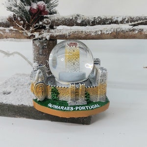 May include: A snow globe featuring a miniature castle with a tower, surrounded by a white snow-like substance. The base of the snow globe is decorated with a green and yellow castle wall and the text "GUIMARAES - PORTUGAL".