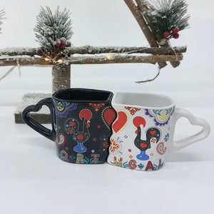 Könnte beinhalten: Zwei Keramikbecher mit herzförmigem Griff. Die Becher sind miteinander verbunden und zeigen ein schwarz-weißes Hahnendesign mit roten Akzenten. Die Becher sind mit portugiesisch inspirierten Mustern verziert.