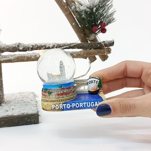 May include: A snow globe featuring a cityscape with a bridge and a boat. The base of the snow globe is blue and white with the text "PORTO-PORTUGAL".