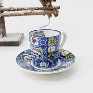 Può includere: Una tazzina da caffè e un piattino in ceramica blu e bianca con un design portoghese. La tazza e il piattino presentano un gallo, un pesce e la parola "Portogallo" in un carattere decorativo.
