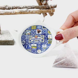 May include: A white ceramic tea infuser with a blue and yellow design featuring Portuguese landmarks and the word "PORTUGAL". The infuser is shaped like a teapot and has a handle. A tea bag is in the background.