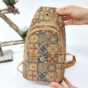 May include: A cork sling backpack featuring a vibrant, geometric tile design. The bag has a zipped main compartment and a smaller front pocket. The strap is light brown. The bag is held by two hands.