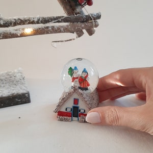 Könnte beinhalten: Eine Schneekugel mit einem kleinen Haus und zwei Figuren darin. Das Haus hat ein rotes Dach und weiße Wände mit dem Wort "MADEIRA" auf der Vorderseite. Die Figuren tragen rote und blaue Kleidung.