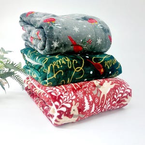 Könnte beinhalten: Ein Stapel von drei gefalteten Decken zum Thema Weihnachten. Die obere Decke ist grau mit roten Vögeln und Schneeflocken. Die mittlere Decke ist grün mit goldenen Buchstaben und einem roten Vogel. Die untere Decke ist rot mit weißen Hirschen und Bäumen.