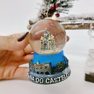 May include: A clear glass snow globe featuring a detailed miniature of a cathedral. The globe sits on a blue base with the words "Viana do Castelo" and a depiction of a stone bridge. The background includes a snowy branch.
