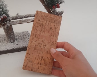 May include: A rectangular cork wallet with a natural, textured surface. The wallet is held in a hand, showcasing its light brown color and stitched edges. The background includes a decorative winter scene.