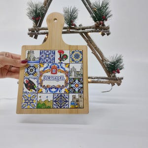 May include: A square wooden cutting board with a handle. The board is decorated with a colorful tile design featuring Portuguese motifs, including a rooster, a coat of arms, and the word "PORTUGAL".