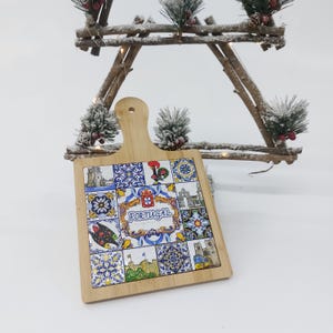 May include: A square wooden cutting board with a handle. The board is decorated with colorful Portuguese tiles featuring traditional designs, including a rooster, fish, and a coat of arms. The word "PORTUGAL" is printed in a gold frame on the board.