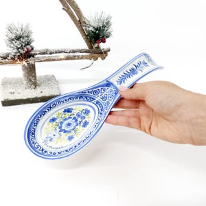 May include: Blue and white ceramic spoon rest with floral design. The spoon rest has an oval shape with a handle. The floral design includes blue flowers and green leaves. The word "Portugal" is written on the spoon rest.