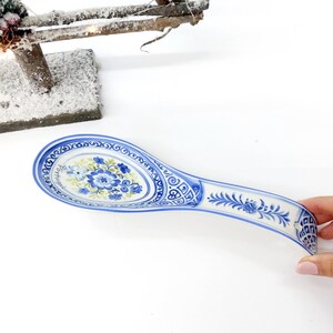 May include: A blue and white ceramic spoon rest with a floral design. The spoon rest has a curved handle and an oval-shaped bowl. The bowl features a central floral motif in blue and green, surrounded by a decorative border. The handle is adorned with floral and geometric patterns.