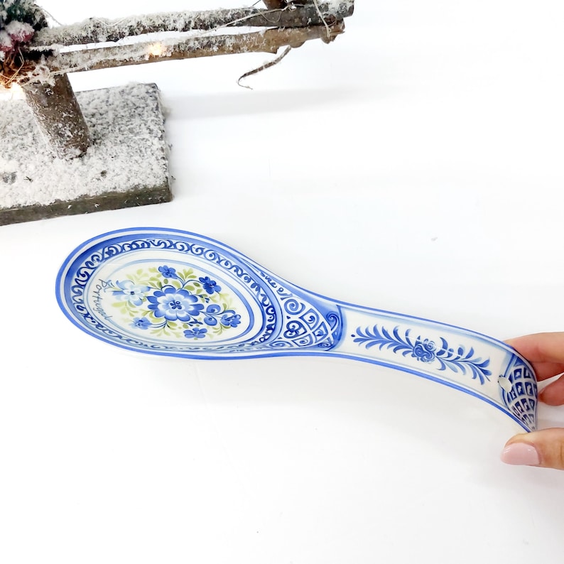 May include: A blue and white ceramic spoon rest with a floral design. The spoon rest has a curved handle and an oval-shaped bowl. The bowl features a central floral motif in blue and green, surrounded by a decorative border. The handle is adorned with floral and geometric patterns.
