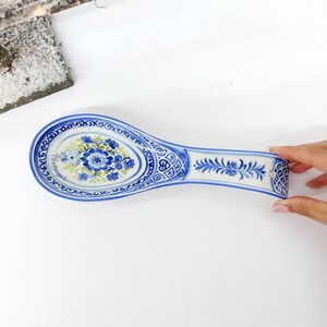 May include: A blue and white ceramic spoon rest with a floral design. The spoon rest has a long, oval shape with a handle. The floral design features blue flowers and green leaves. The word "Portugal" is written on the side.