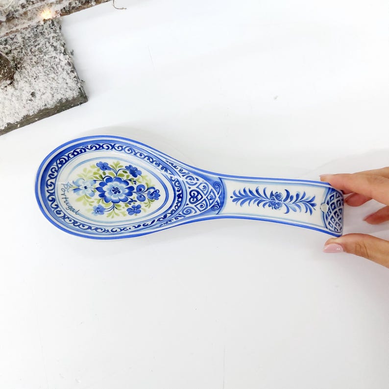 May include: A blue and white ceramic spoon rest with a floral design. The spoon rest has a long, oval shape with a handle. The floral design features blue flowers and green leaves. The word "Portugal" is written on the side.