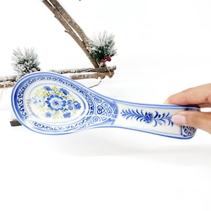 May include: A white ceramic spoon rest with blue floral and scrollwork designs. The spoon rest has a blue rim and handle, with the word "Portugal" written on the side. The spoon rest is held by a person.