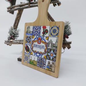 May include: A square wooden cutting board with a handle. The board has a colorful tile design with images of Portuguese landmarks and the word "PORTUGAL" in the center.
