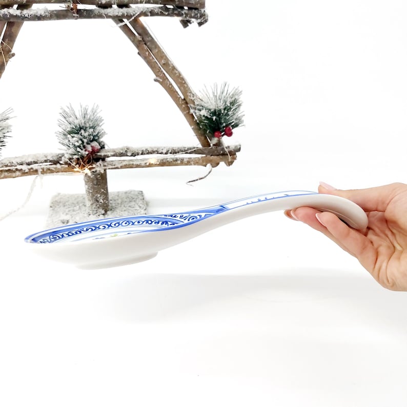 May include: White ceramic spoon with a blue and white decorative pattern along the rim. The spoon has a curved handle and a wide, shallow bowl. The spoon is held against a white background.