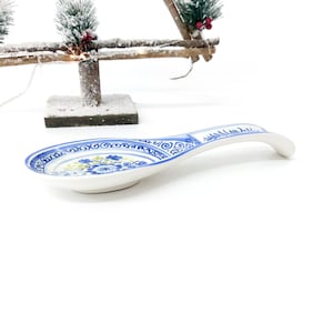 May include: A white ceramic spoon rest with a blue floral design and a curved handle. The spoon rest has a blue border with decorative patterns. The floral design includes blue and yellow flowers. The spoon rest is on a white background.