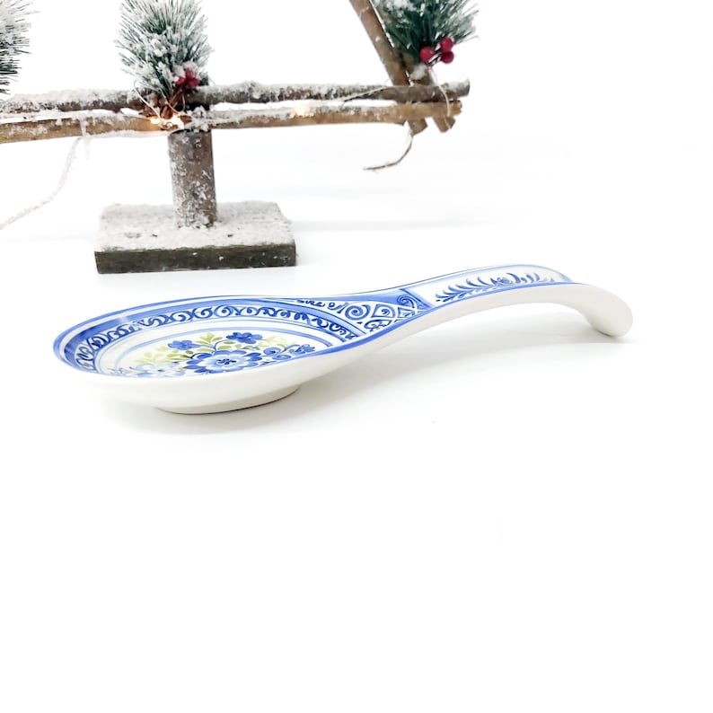 May include: A white ceramic spoon rest with a blue floral design and a curved handle. The spoon rest has a blue border with decorative patterns. The floral design includes blue and yellow flowers. The spoon rest is on a white background.
