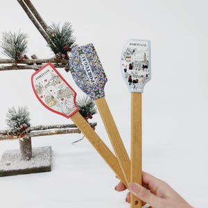 Puede incluir: Tres espátulas de silicona con mango de madera con diferentes diseños que presentan imágenes portuguesas. Las espátulas están impresas con la palabra "Portugal" y presentan una combinación de colores rojo, azul y blanco.