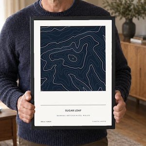 May include: A framed topographic map print featuring white contour lines on a dark navy blue background. The print displays the text SUGAR LOAF, BANNAU BRYCHEINIOG, WALES, 596 m / 1,955 ft, and coordinates 51.8627°N / 3.0579°W in black text on a white border.