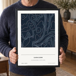 May include: A framed topographic map print of Cairn Gorm, Scotland, featuring white contour lines on a dark blue background. The print includes the text CAIRN GORM, CAIRNGORMS, SCOTLAND, 1,245 m / 4,085 ft, and coordinates 57.1167°N / 3.6442°W.