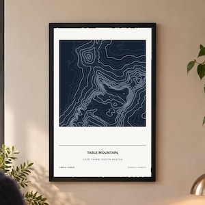 May include: A framed wall art print featuring a dark blue topographic map of Table Mountain in Cape Town, South Africa. The print includes white contour lines, the text Table Mountain, Cape Town, South Africa, 1,085 m / 3,560 ft, and 33.9575°S / 18.4021°E.