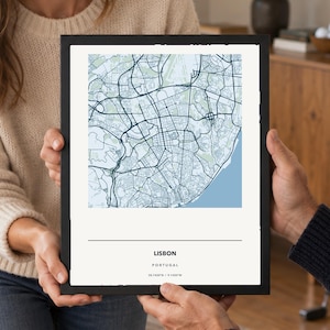 May include: A framed map print of Lisbon, Portugal, featuring a detailed street grid in shades of blue and light green on a white background. The text below the map reads LISBON, PORTUGAL, and the coordinates 38.7420°N / 9.1430°W in black font.