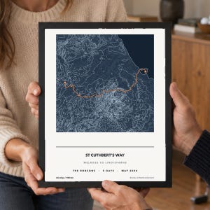 May include: A framed topographic map print titled ST CUTHBERT'S WAY, MELROSE TO LINDISFARNE. The dark blue map features a bright orange route line with markers. Text includes THE ROBSONS, 5 DAYS, MAY 2026, and the distance 62 miles / 100 km.