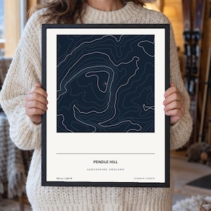 May include: A framed print featuring a dark blue topographic map of Pendle Hill in Lancashire, England. The design includes white contour lines, the text PENDLE HILL, LANCASHIRE, ENGLAND, and the elevation of 557 m / 1,827 ft with coordinates.