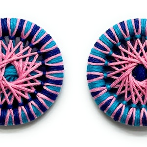 May include: Two round, handmade buttons with a pink and blue striped design. The center of each button is a pink starburst pattern with a blue center.