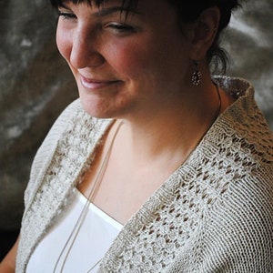 Può includere: Una donna indossa un cardigan in maglia beige chiaro con apertura frontale su un top bianco. Il cardigan presenta un delicato motivo traforato. È visibile una collana d'argento con un pendente verde. La donna sorride.