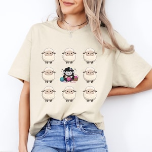 Puede incluir: Una camiseta beige con un gráfico de nueve ovejas blancas y una oveja negra tejiendo con hilo. La oveja negra está rodeada de ovejas blancas.