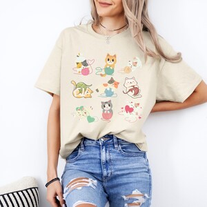 Puede incluir: Una camiseta beige con un estampado gráfico de nueve gatos de dibujos animados jugando con ovillos de lana. Los gatos están en varias poses y colores, incluyendo naranja, blanco, gris y marrón. La camiseta tiene mangas cortas y cuello redondo.