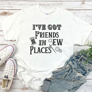 Puede incluir: Camiseta blanca con texto negro que dice "Tengo amigos en lugares de costura". El texto está rodeado de imágenes con temática de costura, incluyendo un carrete de hilo, una aguja y botones.