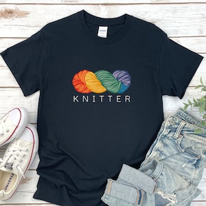 Puede incluir: Camiseta negra con un gr&aacute;fico de ovillo de lana arco&iacute;ris y el texto "KNITTER" impreso en blanco.