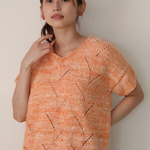 May include: Orange and white knit top with a V-neck and short sleeves. The top features a chevron pattern with small cut-out details. The knitwear is a blend of orange and white threads, creating a textured appearance. The garment is a casual style.