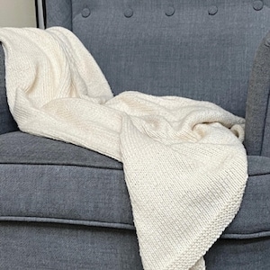 May include: A cream-colored knitted blanket draped over a gray armchair. The blanket has a textured, ribbed pattern and is folded to show its thickness. The armchair has a button-tufted back and a curved design.