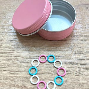 May include: A pink metal tin with a matching lid, slightly ajar, revealing a silver interior. A collection of small, colorful stitch markers in pink, white, and turquoise are arranged in a circle on a wooden surface. The tin is empty.