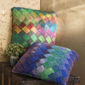 May include: Two colorful knitted throw pillows with a geometric pattern. The pillows are made with a variety of shades of green, blue, purple, and brown yarn. The pillows are sitting on top of a wooden trunk.