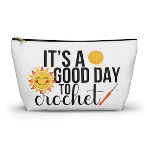 May include: White zippered pouch with the text "IT'S A GOOD DAY TO crochet". The design includes a smiling sun, a ball of yellow yarn, and a crochet hook. The pouch is designed for crochet enthusiasts.